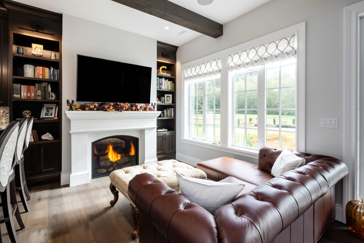 A cozy lounge with a leather sofa, roaring fireplace, large TV, and traditional white vinyl windows.